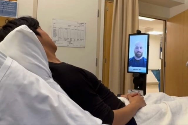 Patient in hospital bed having a video call with a healthcare professional on a screen-mounted device in the hospital room.