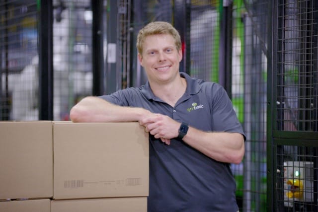 Man in a gray polo shirt leaning on cardboard boxes in a warehouse with industrial shelving in the background