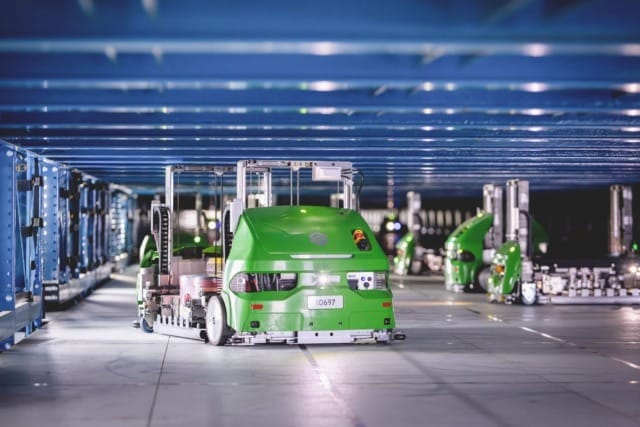 Green autonomous warehouse robots moving under a blue metal framework for automated inventory management.
