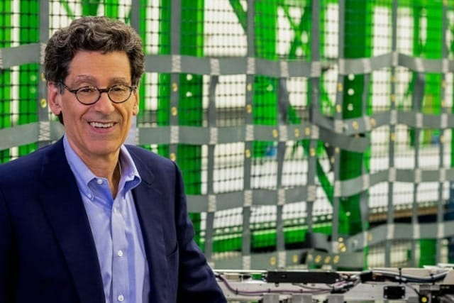 Man in a suit smiling and standing beside industrial machinery with green framework in the background.