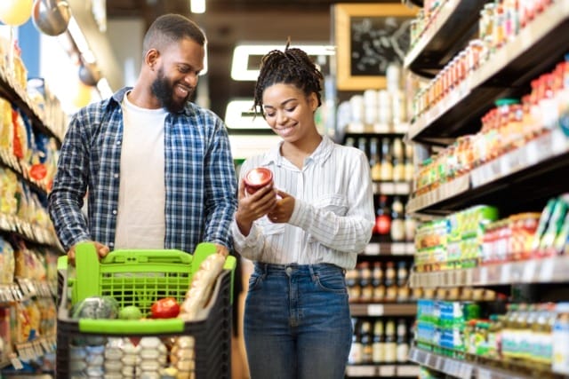 Couple grocery shopping, smiling while examining a product in a supermarket aisle filled with various items.