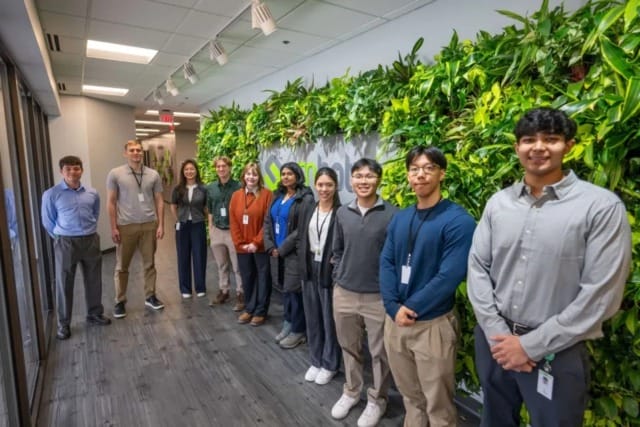 Group of people standing in an office hallway with green plant wall, smiling and wearing ID badges.