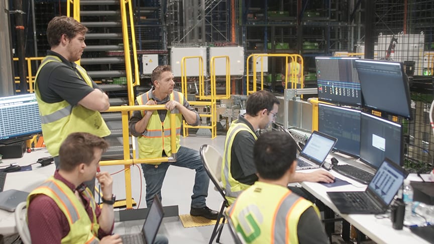 Warehouse workers in safety vests collaborate at a workstation with multiple computer screens.