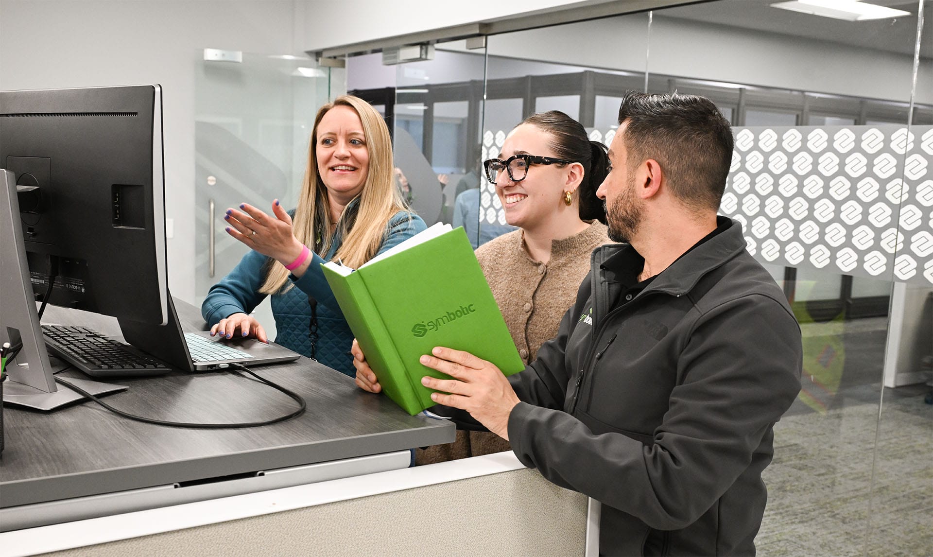 Three people smiling and discussing at a computer in an office setting with a green book titled "Symbolic" in hand.